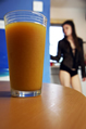 A glass of orange juice on a table with a girl in a sheer blouse in the background