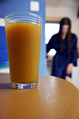 A glass of orange juice on a table with a girl in a silky blue wrapover robe in the background