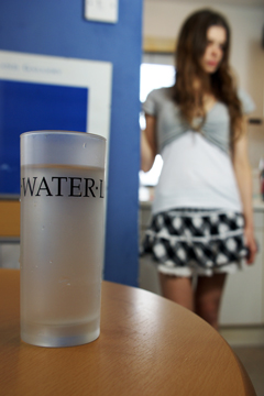A glass of water on a table with a girl in a miniskirt in the background