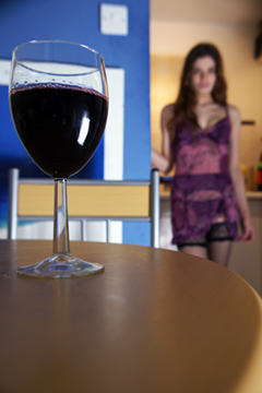 A glass of red wine on a table with a girl in a sheer slip and lingerie in the background