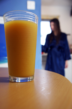 A glass of orange juice on a table with a girl in a silky blue wrapover robe in the background