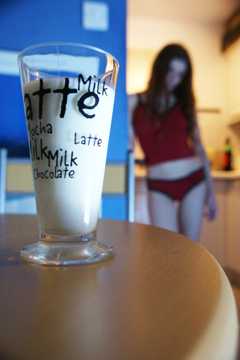 A glass of milk on a table with a girl in a vest top and knickers in the background