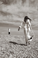 Girl in white lace carries strappy sandals and walks away towards lighthouse