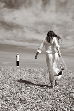Girl in white lace carries strappy sandals and walks away towards lighthouse
