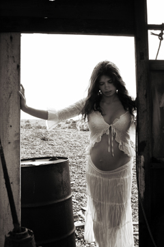 A pregnant woman in white lace stands illuminated in the doorway to an old wooden hut
